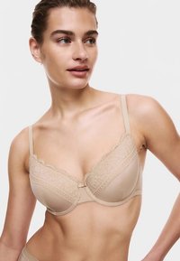 Béžová podprsenka s hladkou látkovou základnou, krajkovým lemem podél okrajů a nastavitelnými ramínky. Zapínání vpředu na háček s dekorativní mašlí.