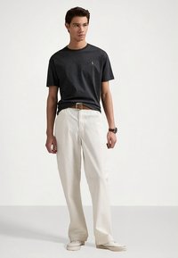 Tamsiai pilka medvilninė marškinėliai su mažu logotipu, derinami su laisvo kirpimo baltais kelnėmis ir šviesios spalvos avalyne, papildyta rudu diržu.