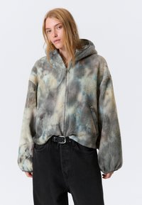 Fleecejakke med hette, med et tie-dye mønster i grå, blå og kremfarger. Glidelås foran, tettsittende mansjetter og avslappet passform.