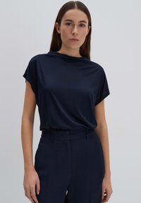 Top blu navy a maniche corte con scollatura drappeggiata, realizzato in tessuto morbido, abbinato a pantaloni blu navy sartoriali, creando un look coerente.