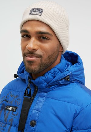 Padded blue jacket with a zip front and black hardware, featuring logo accents. Cream ribbed beanie on the head. Smooth, shiny fabric.