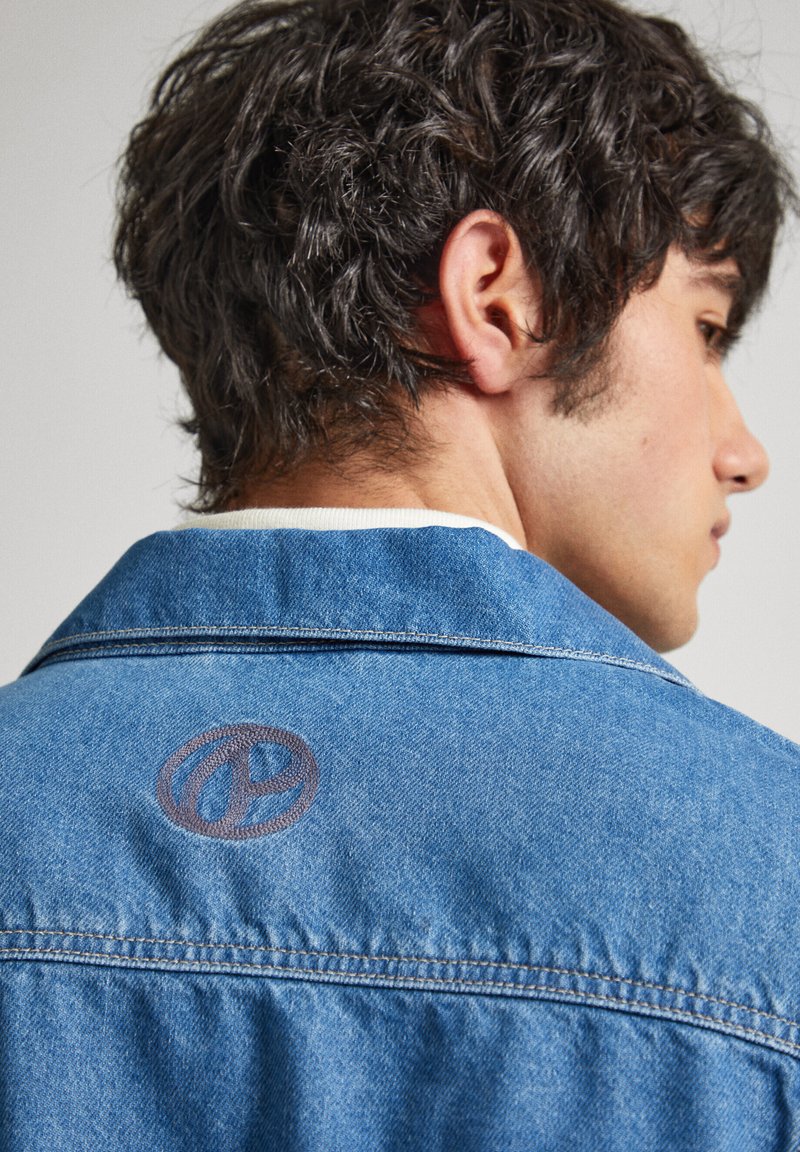 Chaqueta de mezclilla azul con collar puntiagudo, costuras visibles y un detalle de logo circular marrón en la parte superior de la espalda. Textura fina visible.