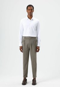 Chemise blanche à manches longues avec fermeture boutonnée, associée à un pantalon gris taille haute avec plis et accents de boutons, complétée par des mocassins marron.