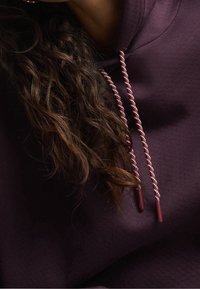 Felpa borgogna con tessuto testurizzato, caratterizzata da lacci attorcigliati rosa e bianchi con punte in plastica, completata da capelli mossi in primo piano.