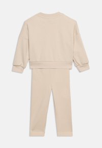 Bež komplet dolge majice z dolgimi rokavi in ujemajočih hlač, prikazan od zadaj, z rebrastimi manšetami in spodnjim robom na majici.