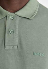 Grønn polo skjorte laget av teksturert stoff med klassisk krage, og har tre matchende knapper samt en logo nederst til høyre.