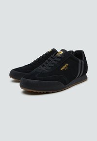 Scarpe sportive in suede nero con suola in gomma, caratterizzate da un logo dorato e strisce nere sui lati, e un classico design con lacci.