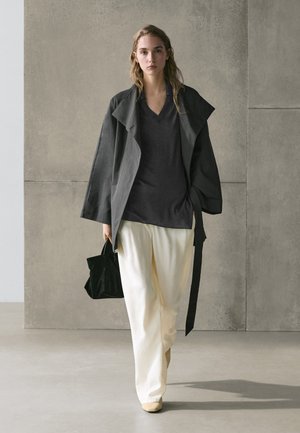 Young woman walking indoors wearing a dark gray oversized jacket, black top, white wide pants, beige shoes, and carrying a black handbag.