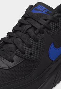 Κοντινό πλάνο μαύρου αθλητικού παπουτσιού Nike με μπλε λογότυπο στο πλάι και στη γλώσσα, με υφασμένα κορδόνια και διάτρητες λεπτομέρειες.