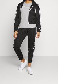 Svart cropped jacka med vita ränder, grå t-shirt med logotyp, svarta byxor med sidostripes och vita sneakers, allt i en sportig stil.