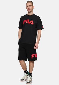 Sort kortærmet Fila T-shirt med stort rødt logo, parret med sorte shorts med rødt Fila-logo, suppleret med hvide og sorte sneakers.