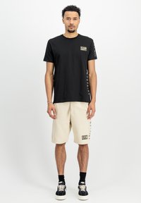 Sort bomulds t-shirt med camo-patch og lodret tekst på ærmet, parret med beige shorts med en struktureret logo-patch.