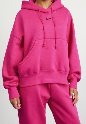 Person trägt knallpinkes oversized Nike Hoodie und passende Jogginghose mit entspannten Händen an den Seiten vor schlichtem Hintergrund.