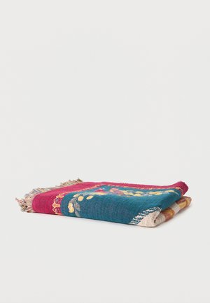 Folded colorful woven blanket with red, teal, and beige sections, floral patterns, and fringed edges on a white background.