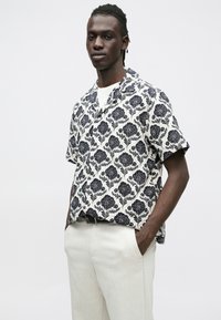 Koszula z krótkim rękawem w kremowym kolorze z granatowym wzorem kwiatowym. Noszona z jasnymi spodniami, prezentująca luźny krój i kołnierzyk.