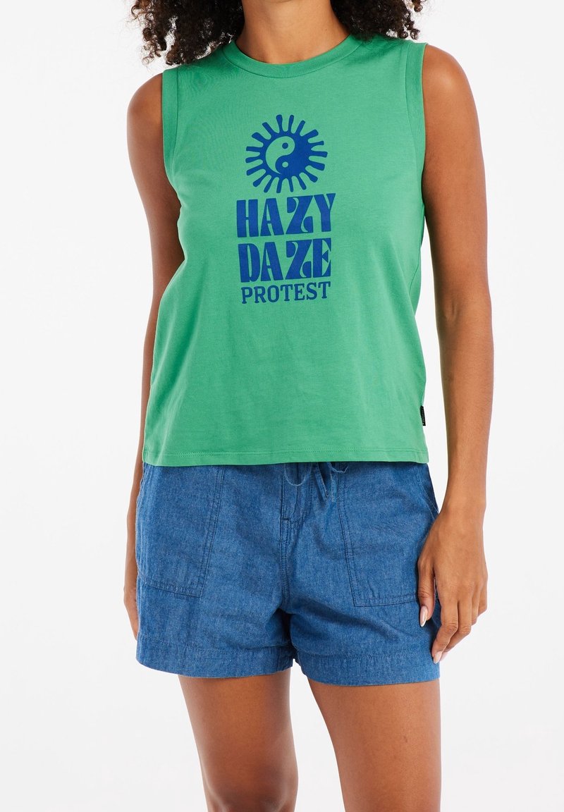 Femme portant un haut sans manches vert avec un texte bleu « Hazy Daze Protest » et un graphisme soleil yin-yang, associé à un short en jean bleu.