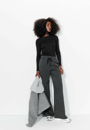 Haut à manches longues noir, pantalon à cordon grisé à carreaux, tenant une couverture en tricot grise. Des baskets argentées complètent la tenue, le tout sur un fond blanc.