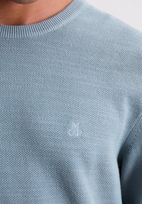 Pull en tricot bleu clair avec un col ras du cou côtelé et un petit logo brodé sur la poitrine, mettant en avant un tissu texturé.
