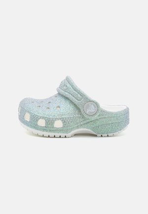 Lyseblåt glitrende slip-on clog med ventilationshuller og en bagrem med en cirkulær logo på en hvid sål.