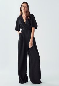 Ženska stoji s roko v žepu, oblečena v črno široko hlačno enodelno obleko s kratkimi lepršavimi rokavi in V-izrezom na svetli podlagi.