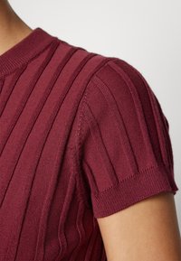 Gros plan d'une personne portant un haut en maille côtelée à manches courtes, de couleur bordeaux profond avec des rayures texturées verticales.