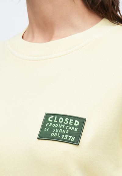 Sweat-shirt jaune clair avec un col rond et un patch vert brodé portant l'inscription "CLOSED PRODUTTORE DI JEANS DAL 1978."
