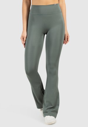 Donna che indossa leggings svasati a vita alta di colore verde oliva e sneakers bianche, in piedi con le mani rilassate ai fianchi.
