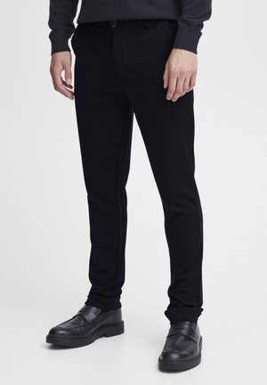 Pantalones ajustados negros hechos de una tela suave, con un diseño entallado y bolsillos laterales, combinados con zapatos negros sin cordones.