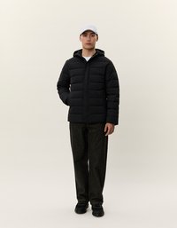 Sort pufferjakke med lynlås foran, hætte og quiltet design. Båret med mørke denim-bukser og sorte sko, kompletteret af en hvid kasket.