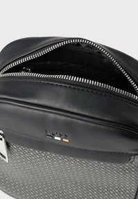 Czarna skórzana torba z srebrnym suwakiem, czarno-szarym wzorem o fakturze oraz logo z białymi, czarnymi i pomarańczowymi akcentami na froncie.