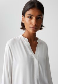 Vrouw met kort donker haar, gekleed in een witte blouse met lange mouwen en een V-hals, met een neutrale uitdrukking, tegen een effen lichte achtergrond.