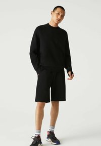 Sudadera negra con cuello redondo y mangas largas, combinada con pantalones cortos negros. Llevada con zapatillas deportivas negras, rojas y azules.