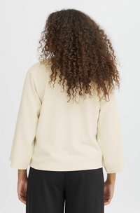 Suéter de color beige claro con textura de canalé, mangas anchas de tres cuartos y dobladillo redondeado. Diseño minimalista, combinado con pantalones negros.