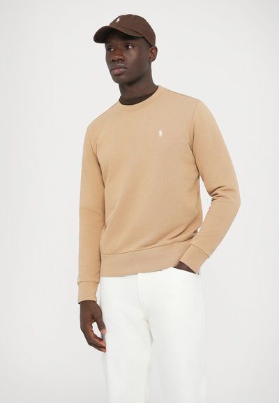 Bež pulover z rebrastim okroglim izrezom in manšetami, z majhnim vezenim logotipom. Nošen z belimi hlačami in rjavo kapo.