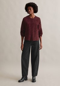 Burgundowa, teksturowana koszula z długim rękawem, zestawiona z luźnymi czarnymi dżinsami i masywnymi czarnymi butami. Neutralne tło.