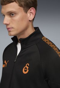 Schwarze Zip-Jacke mit orangefarbenem Logo und Akzenten, mit hohem Kragen und strukturierten Streifen entlang der Schultern. Weiche Stoffoberfläche.