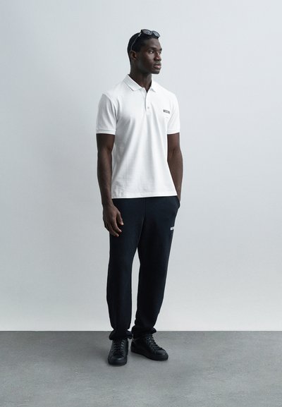 Άνδρας που φοράει λευκό πόλο μπλουζάκι, μαύρο παντελόνι τζόγκινγκ, μαύρα αθλητικά παπούτσια και γυαλιά ηλίου στο κεφάλι, στέκεται μπροστά από ένα απλό ανοιχτό γκρίζο φόντο.