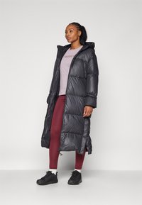 Lång svart puffjacka med huva, quiltat mönster, sidofickor, parat med en ljuslila skjorta och vinröda leggings, samt svarta sneakers.