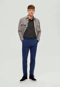 Camisa cinza de botões sobre um pólo escuro, calças azuis e ténis pretos. Conjunto casual com cores e texturas contrastantes.