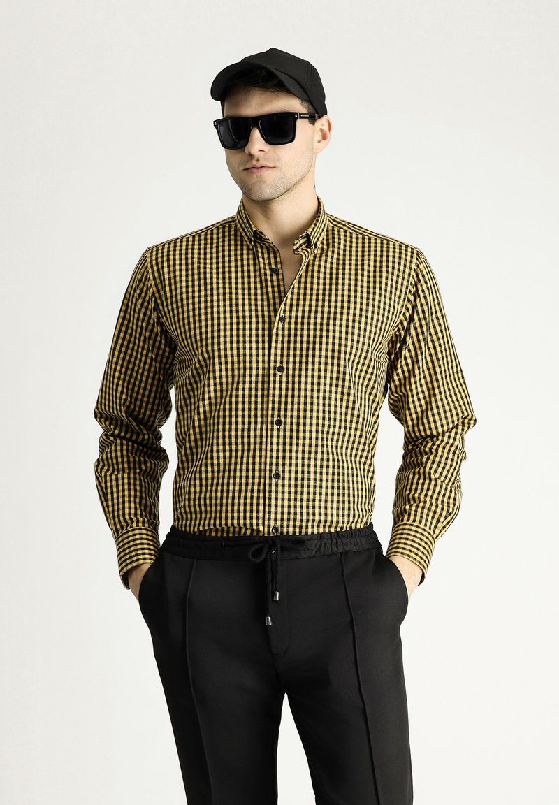 Camisa a cuadros amarilla y negra con mangas largas, abertura en la parte delantera con botones y un collar. Llevada con pantalones negros y complementada con gafas de sol y una gorra.