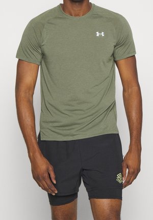 Mann trägt olivgrünes kurzärmeliges Sportshirt und schwarze, mehrlagige Sportshorts, steht vor einfarbigem Hintergrund.