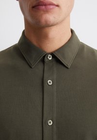 Olive green polo shirt with a structured collar, featuring a two-button placket and textured fabric. Visible stitching accents near the buttons.
