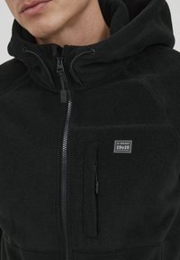 Mustat fleece-takki, jossa on täyspitkä vetoketju, säädettävä huppu ja sivutasku, varustettu neliön muotoisella logolla. Pehmeä pinta ja lämmin muotoilu.