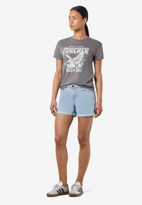 Camiseta gráfica gris con diseño de águila, combinada con pantalones cortos de denim azul claro. Ambos tienen un corte casual, complementados con zapatillas blancas y grises.