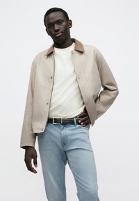 Giacca beige chiaro con colletto in velluto a coste marrone, t-shirt bianca, jeans blu e cintura nera; tessuto liscio con bottoni a pressione.