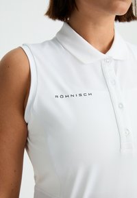 Biała bezrękawnikowa koszulka polo z guzikiem przy kołnierzyku oraz czarnym logo "Röhnisch" na piersi, wykonana z gładkiego, lekko teksturowanego materiału.