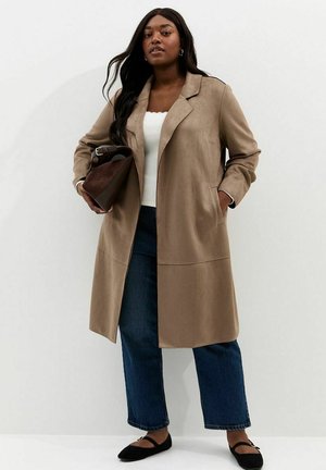 Frau mit langen dunklen Haaren, die einen beigen Mantel, ein weißes Oberteil, blaue Jeans, schwarze Ballerinas trägt und eine braune Handtasche hält, steht vor einer weißen Wand.