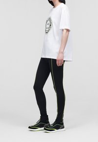 Witte katoenen T-shirt met een ronde zwarte logo. Zwarte legging met groene zijstrepen. Zwarte sneakers met groene accenten.