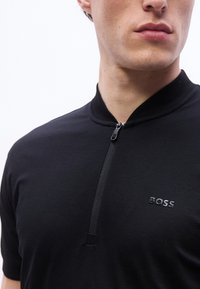 Άνδρας που φορεί μαύρο κοντομάνικο πουκάμισο με γιακά φερμουάρ και το λογότυπο "BOSS" στο αριστερό στήθος, εμφανιζόμενος από τον λαιμό έως το μέσο του κορμού.