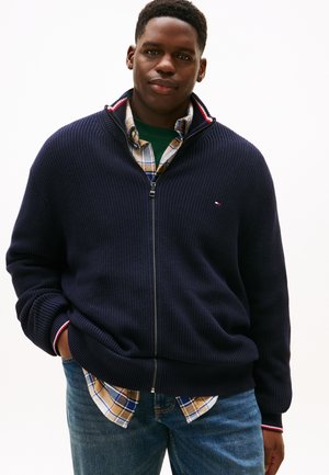 Ung mand iført en marineblå zip-up sweater over en ternet skjorte og grøn t-shirt, med hænderne i lommerne, stående mod en ensfarvet hvid baggrund.